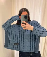 Sweater de punto con diseño de trenzas verticales en color azul piedra. Presenta un corte crop, cuello redondo, mangas largas con puños acanalados y paneles laterales en tejido liso. Incluye una etiqueta decorativa en la parte frontal inferior.