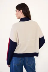 Sweater de punto acanalado con cuello alto y cierre frontal parcial. Presenta un diseño tricolor en bloques con cuerpo color crema, cuello y puños azul marino, y detalles en rojo en las mangas. Incluye una pequeña etiqueta con logo en el pecho.