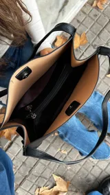 Cartera de mano tipo tote en color negro, confeccionada en cuero con textura granulada. Presenta un diseño minimalista con doble asa superior y un compartimento interior en tono contrastante.