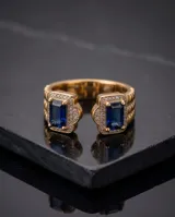 Anillo abierto de bronce con baño de oro, adornado con dos piedras rectangulares de color azul zafiro rodeadas de circonias brillantes y detalles texturizados en los laterales.