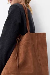 Bolso tipo tote de gran tamaño, confeccionado en gamuza color tostado. Presenta un diseño minimalista con doble asa larga para llevar al hombro y un cierre mediante cordón ajustable en la parte superior.