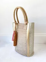 Bolso matero color beige con textura tipo piel de cocodrilo y detalles dorados. Tiene doble asa de mano y un dije decorativo con flecos rosas y marrones.