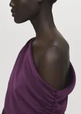 Vestido midi de satén color violeta oscuro, con diseño drapeado en el escote y un hombro al descubierto, de manga corta caída y bajo asimétrico.