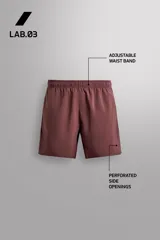 Pantalones cortos deportivos color marrón rojizo, confeccionados en tejido técnico ligero y elástico. Presentan cintura elástica ajustable con cordón interior, bolsillos frontales y un bolsillo trasero con cierre de cremallera. Los laterales tienen perforaciones para transpirabilidad y aberturas laterales en el bajo.