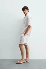 Camisa de corte relajado confeccionada en algodón, con cuello bowling y manga corta. Presenta un bolsillo en el pecho con vivo a contraste y bordados decorativos en el frente. Cierre frontal mediante botones.
