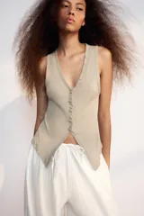 Top de algodón color beige con escote en V, sin mangas y cierre frontal con botones. Presenta un diseño asimétrico en el bajo con abertura delantera.