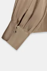 Blusa de tejido satinado color bordó, con cuello subido drapeado y manga larga con puños. Presenta detalle de pliegues frontales y cierre con botones laterales en el cuello.
