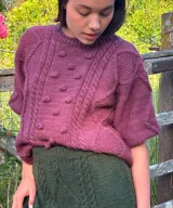 Sweater de punto color bordó, de manga corta, con diseño amplio y relajado. Presenta detalles de trenzas, rombos y bodoques en relieve en la parte frontal y mangas.