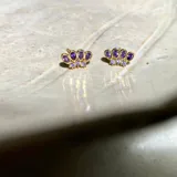 Par de aros dorados con forma de corona, con cinco circonias púrpuras en forma de gota y tres circonias blancas pequeñas en la base. Fabricados en plata 925 con acabado en oro de 18k.