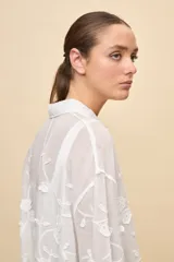 Camisa blanca de calce relajado, con cuello clásico, mangas 3/4 y cierre frontal de botones. Presenta bordados florales en relieve y detalles de crochet calado en los puños y el ruedo.