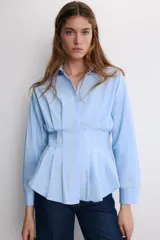 Camisa blanca de popelín con cuello solapa y manga larga, cintura entallada con pliegues y cierre frontal con botones ocultos bajo solapa.