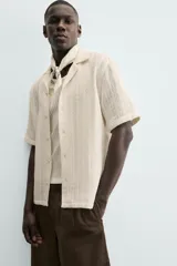 Camisa de calce relajado confeccionada en tejido con algodón de color beige. Presenta cuello bowling, manga corta, aberturas laterales en el bajo y cierre frontal con botones.