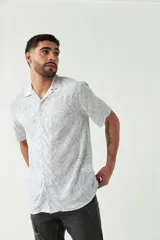 Camisa de manga corta con cuello tipo cubano, confeccionada en viscosa con estampado de hojas en tonos azules sobre fondo blanco. Presenta cierre frontal con botones y corte slim.
