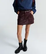 Short pollera de tiro medio confeccionado en poliuretano con acabado símil cuero. Presenta diseño de corte recto, cierre frontal con cremallera, trabillas para cinturón y dos bolsillos delanteros con solapa.
