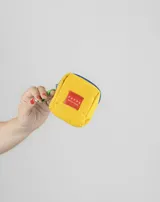 Monedero pequeño de tela color beige con cierre azul y tira de agarre color verde. Incluye un parche rectangular rojo con logo y gancho metálico para colgar.
