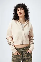 Canguro cropped de color beige claro, con cierre frontal y capucha. Presenta un estampado abstracto en color marrón oscuro en ambas mangas.