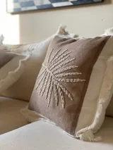 Almohadón cuadrado color marrón claro con bordado de hilo color crema con diseño de rayos de sol. Tiene borde blanco con flecos.