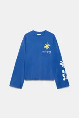 Remera oversize de manga larga color azul, con efecto lavado, cuello redondo y detalle de estampación inspirada en Henri Matisse con una estrella amarilla en el pecho y formas abstractas blancas en la manga.