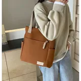 Cartera tipo tote de lona color marrón con asas de mano en color beige. Cuenta con un bolsillo frontal, bolsillos laterales y un detalle de tirador de cierre en cordón blanco.