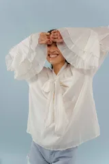 Blusa de algodón color blanco, con cuello alto y lazo frontal para atar. Presenta mangas largas con volados en los puños y un diseño holgado y romántico.