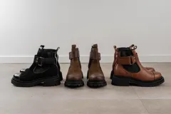 Bota tipo Chelsea de cuero marrón con suela track negra de goma. Presenta paneles elásticos laterales y una costura reforzada en la puntera. Incluye una correa de cuero desmontable con hebilla metálica.