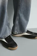 Zapatilla de estilo retro con cordones, confeccionada en una combinación de piezas de cuero sintético negro y detalles en blanco, con suela dentada de goma en tono caramelo.
