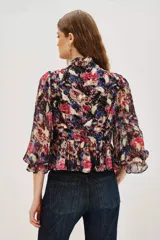 Camisa de manga larga con estampado floral y detalles en hilo metalizado. Presenta cuello alto con volados, cierre frontal con botones y puños con volados. El diseño incluye un corte entallado en la cintura con un ligero peplum.