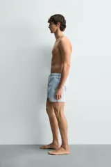 Bañador de hombre de largo medio, color celeste claro con rayas blancas horizontales. Presenta cintura elástica ajustable con cordón, bolsillos frontales y bolsillos traseros de plastrón. Incluye forro interior.