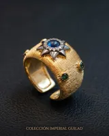 Anillo ancho de bronce con baño de oro, diseño texturizado, estrella central decorada con circonias y piedra azul, y pequeñas piedras verdes en los laterales.