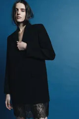 Vestido tipo blazer negro, de corte recto y largo medio, con cuello solapa, manga larga con abertura y botones, y bolsillos con solapa en el delantero. Presenta un bajo con aplicación de encaje negro a tono.