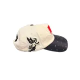 Gorro con visera de color beige y visera negra, con bordado frontal en relieve rojo y detalles de strass distribuidos por toda la prenda.