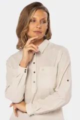 Camisa de lino color beige, manga larga con presilla para sujetar el puño enrollado, cuello camisero y bolsillo de parche en el pecho. Presenta un semi-abertura frontal con botones metálicos.