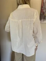 Camisa de manga larga en color beige, con cuello clásico y cierre frontal de botones. Presenta un delicado aplique de encaje tipo crunie en la parte superior delantera, aportando textura y un detalle artesanal al diseño.