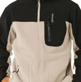 Canguro con cierre frontal de la línea Anti Series de Rip Curl. Presenta un diseño de bloques de color en beige y negro, cuello alto, bolsillos laterales y un bolsillo vertical con cierre en el pecho que incluye el logo de la marca.