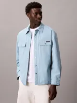Sobrecamisa de algodón de corte regular, color celeste, con cierre frontal de botones a presión, bolsillos frontales con solapa y puños abotonados. Presenta un parche con el logo de la marca en el bolsillo superior izquierdo.