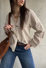 Camisa beige de inspiración boho con detalles bordados en mangas y puños, cuello en V con botones frontales y mangas largas abullonadas.