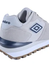 Championes urbanos Umbro de diseño deportivo, confeccionados en una combinación de gamuza y malla en tonos beige y gris claro. Presentan el logo de la marca en el lateral, una entresuela blanca con refuerzo azul oscuro en el talón y una suela dentada en color marrón.