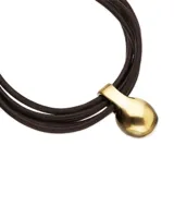 Pulsera de cuero negro con tres tiras, cierre de mosquetón dorado y un dije central en forma de gota, bañado en oro 18K.