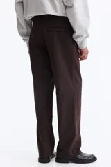 Pantalón sastrero color marrón oscuro, de corte relajado y piernas con pliegues, confeccionado en mezcla de algodón y lyocell. Cuenta con cierre y botón, bolsillos laterales y bolsillos traseros insertados con botón oculto.