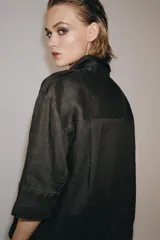 Camisa de lino color marrón oscuro con cuello solapa, manga por debajo del codo y cierre frontal con botones ocultos por solapa. Presenta bolsillos de plastrón con solapa en el pecho y trabillas en los hombros.