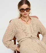 Blusa tipo bata de encaje texturizado color beige, con escote en V, mangas largas y volados aplicados en el escote. Presenta cierre frontal con botones y lazo de ajuste en la cintura.