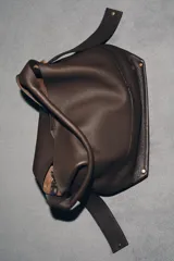 Bolso tipo saco de gran tamaño en cuero marrón oscuro, con textura granulada. Presenta un asa de hombro ancha y detalles metálicos dorados en la unión del asa. Incluye un bolsillo interior extraíble y cierre mediante botón imantado.