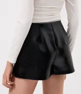 Short pollera de tiro alto confeccionado en poliuretano con acabado que imita el cuero. Presenta un diseño cruzado tipo falda con cinturón integrado de hebilla metálica y cierre oculto.