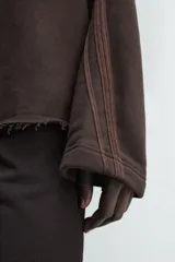 Sudadera de corte boxy fit en color marrón oscuro, con capucha, mangas largas y bandas laterales a contraste. Presenta un diseño de bajo con acabado irregular deshilachado.