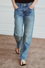 Jeans de tiro medio con corte recto y largo completo. Presentan un diseño de cinco bolsillos, cierre frontal con cremallera y botón, y un acabado con efecto lavado. Incluyen detalles de roturas y parches decorativos en la parte delantera.