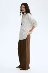 Pantalón sastrero de corte ancho, color marrón, con pinzas frontales, trabillas para cinturón y raya marcada en las piernas.