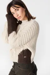 Sweater de punto con diseño de bloques de color en tonos crema y marrón. Presenta cuello redondo, mangas largas y una silueta holgada con detalle de placa metálica decorativa en el lateral.
