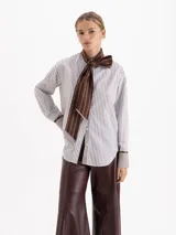 Camisa de poplín con diseño de rayas verticales en tonos blanco y marrón. Presenta cuello con tiras largas integradas para anudar, mangas largas con puños anchos en contraste y cierre frontal con botones.