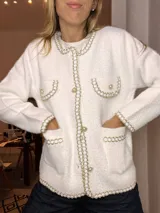 Saco tipo chaqueta de punto texturizado en color blanco, con diseño inspirado en el estilo tweed. Presenta bordes decorados con una trenza metálica dorada y plateada, cierre frontal con botones perlados, bolsillos frontales y detalles de solapas falsas en el pecho.