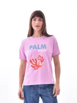 Remera de manga corta color rosa claro con cuello redondo. Presenta un estampado frontal con el texto "PALM BEACH" en azul y un diseño gráfico abstracto de coral en naranja.